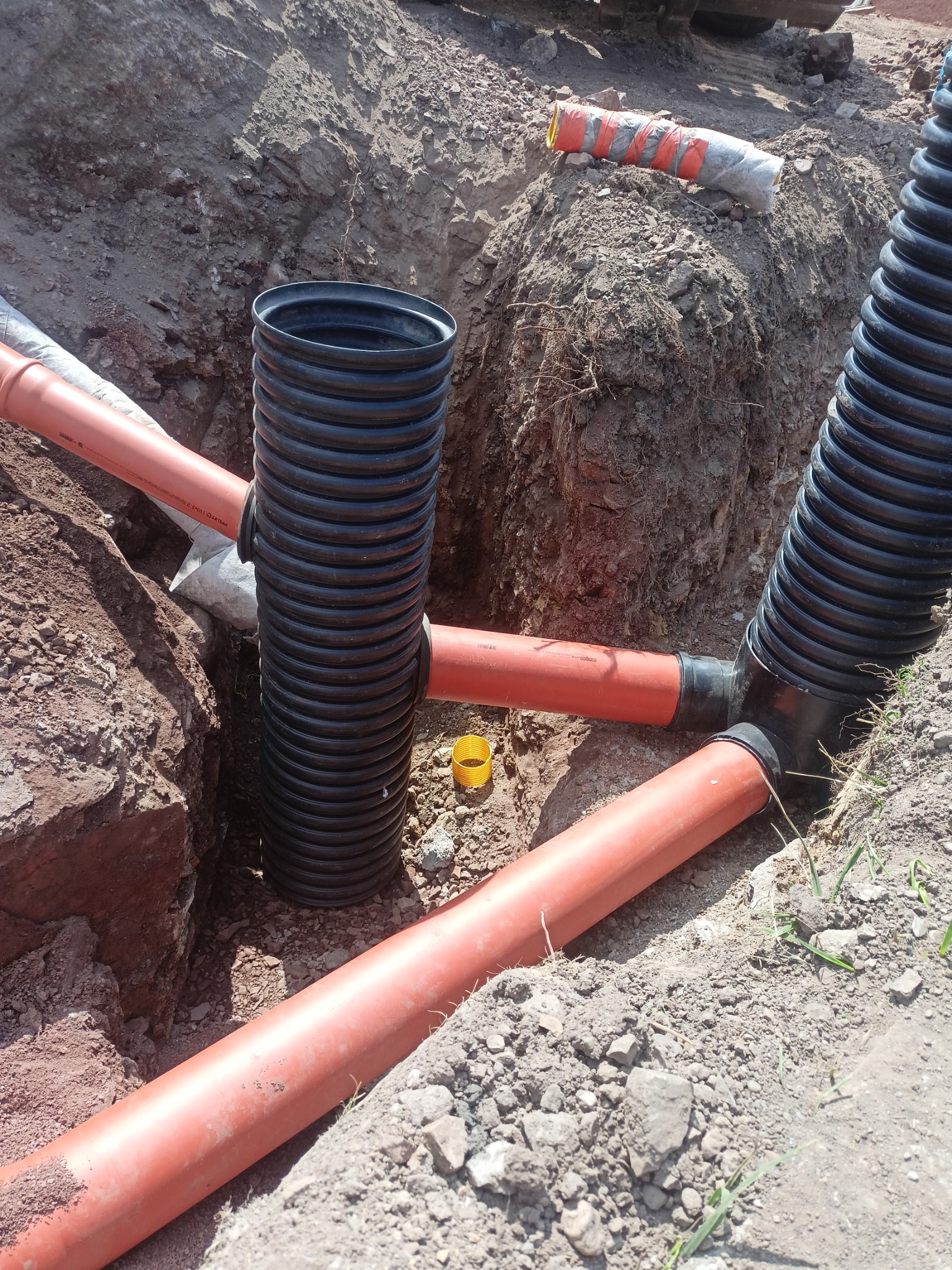 Instalacja podziemna: czarna karbowana rura pionowa połączona z pomarańczowymi rurami w wykopie ziemnym.