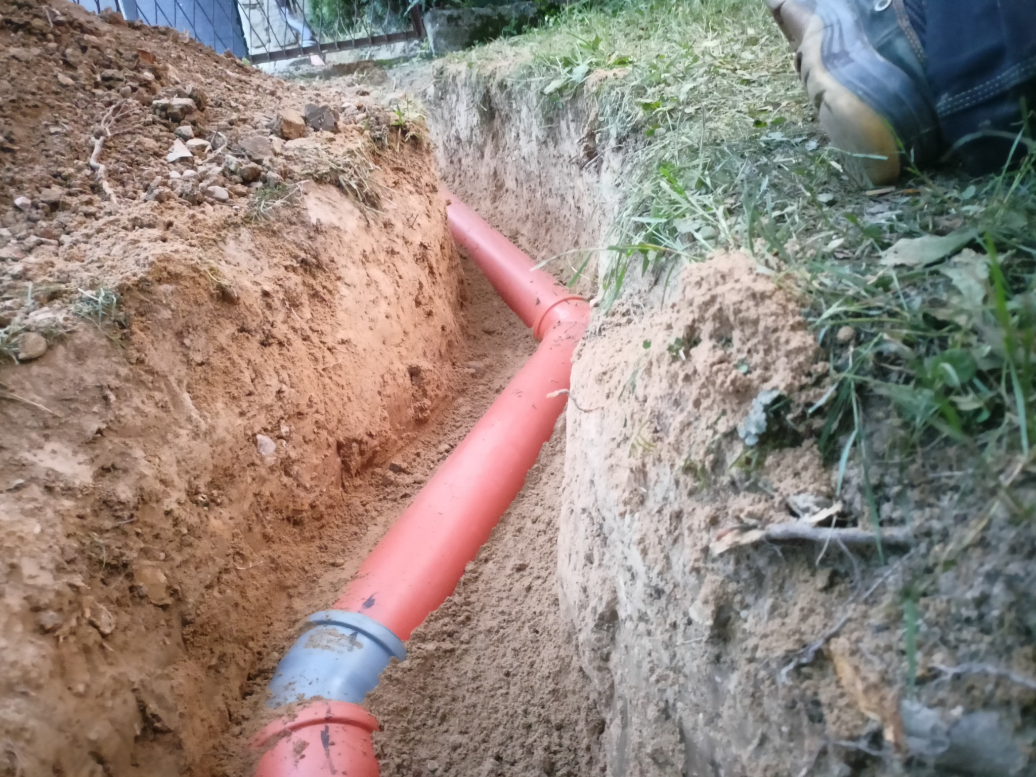Pomarańczowe rury kanalizacyjne z szarymi złączkami ułożone w wykopie w ziemi, widoczne z góry, obok but robotnika.