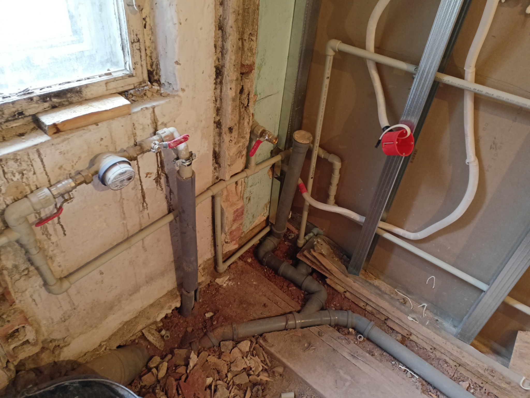 Fragment instalacji hydraulicznej w trakcie przeróbki: szare rury PCV, metalowe profile do zabudowy, fragment okna z widocznymi śladami wilgoci i zniszczeń na ścianie.