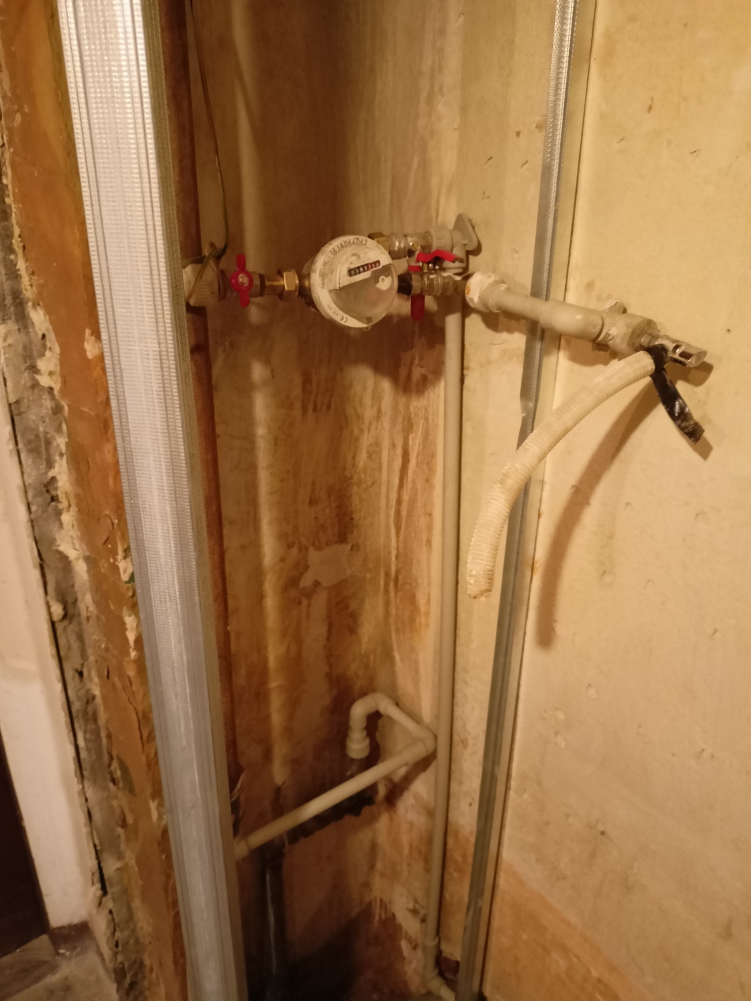 Zamontowany licznik wody z zaworami odcinającymi, rurami i fragmentem stelażu pod zabudowę w surowym wnętrzu.