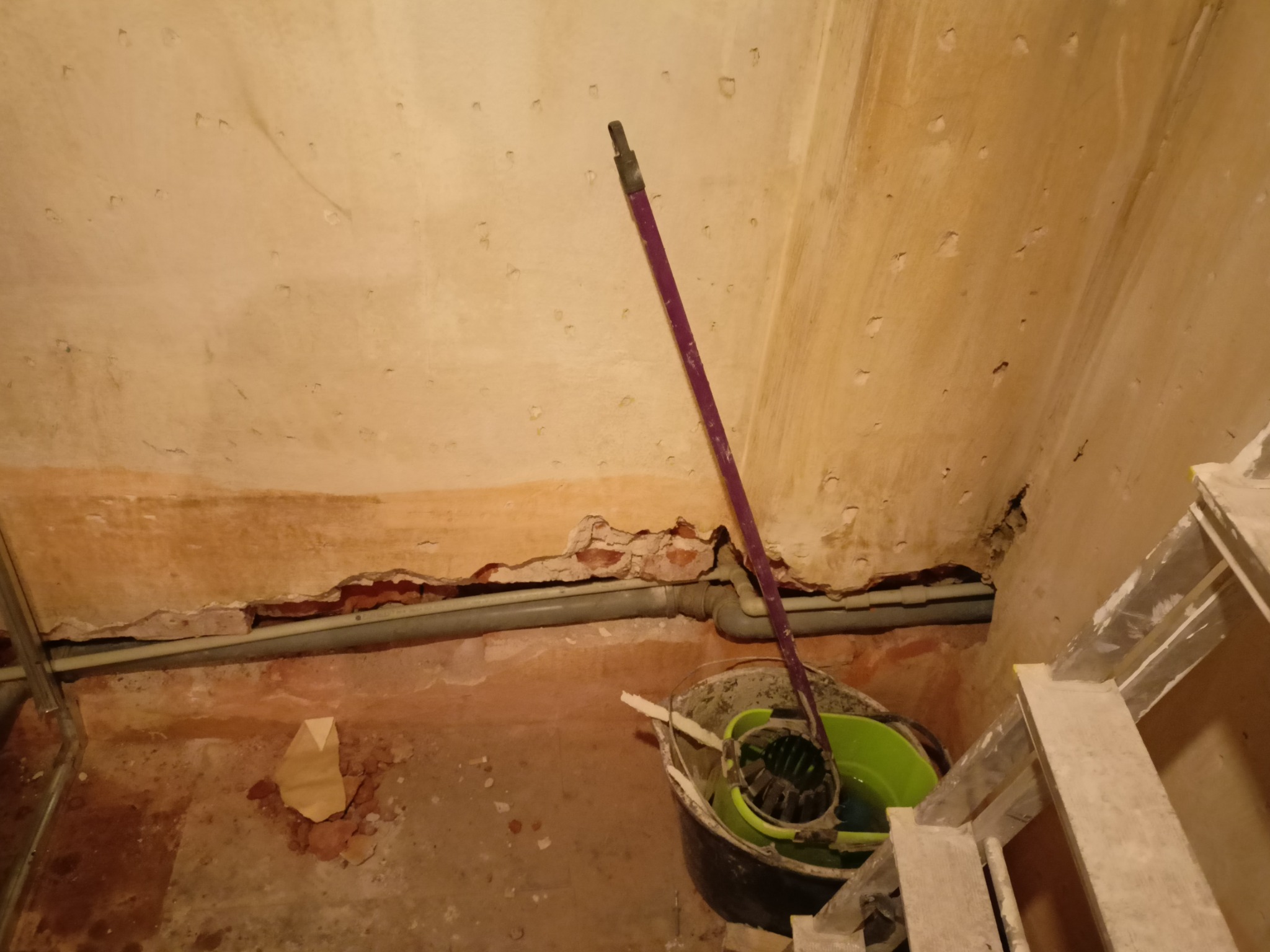 Fragment wnętrza w trakcie remontu: odsłonięta cegła, rura hydrauliczna, wiadro z mopem i drabina w rogu pomieszczenia.