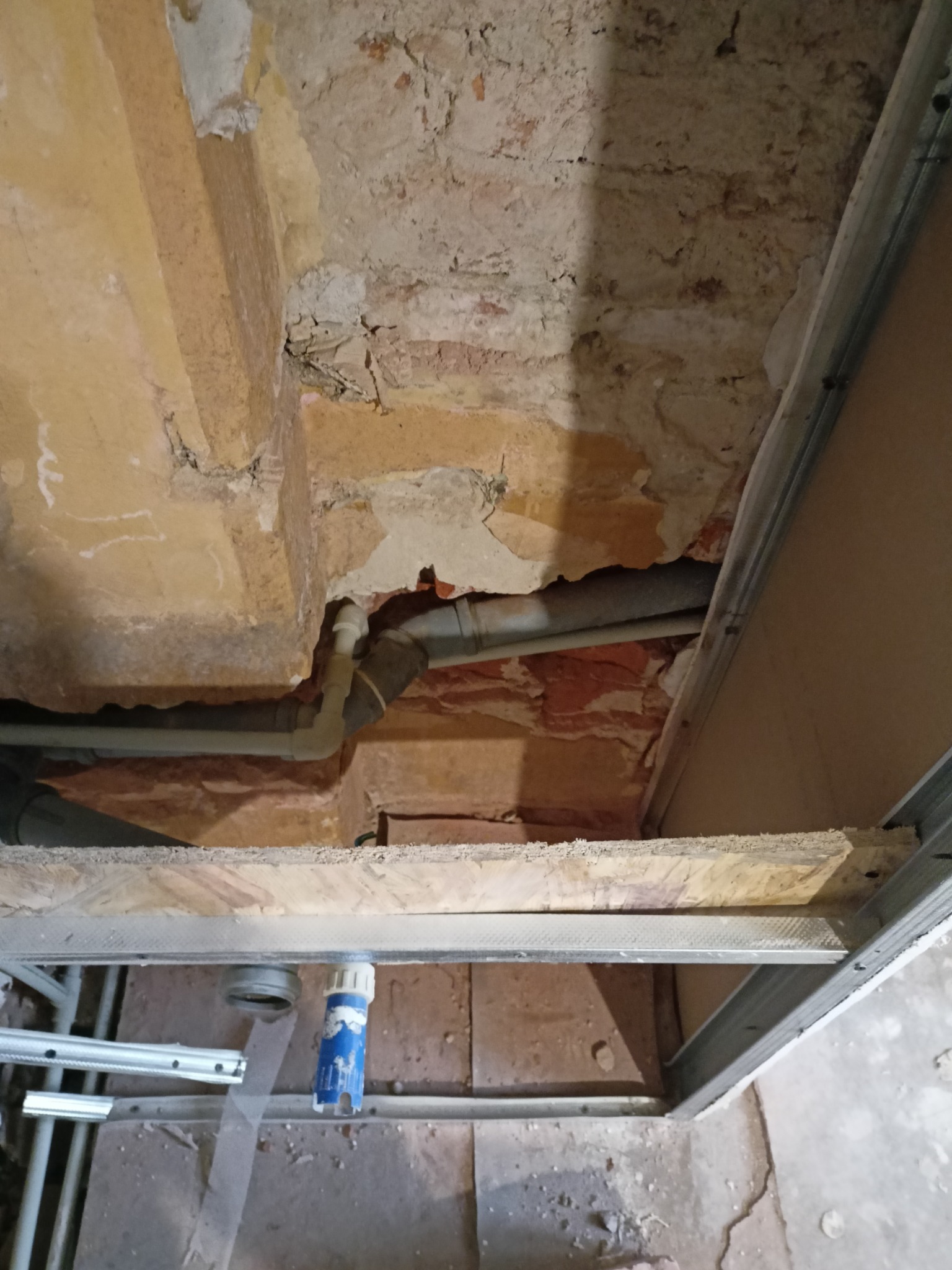 Odsłonięta instalacja hydrauliczna pod stropem z widocznymi rurami, elementami konstrukcji sufitu podwieszanego i fragmentami starej ceglanej konstrukcji.