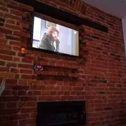 PAMIREX REMONTY I WYKONCZENIA PAWEŁ HAWRO - Telewizor wmurowany w ścianę z czerwonej cegły nad czarnym kominkiem, widoczne niedociągnięcia w wykończeniu ściany wokół telewizora i kabli.
