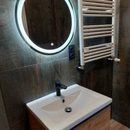 PAMIREX REMONTY I WYKONCZENIA PAWEŁ HAWRO - Nowoczesna łazienka z okrągłym lustrem podświetlanym LED, białą umywalką na drewnianej szafce i grzejnikiem drabinkowym na tle ciemnych płytek imitujących drewno.