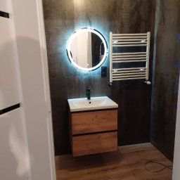 PAMIREX REMONTY I WYKONCZENIA PAWEŁ HAWRO - Nowoczesna łazienka z wiszącą szafką z umywalką, okrągłym lustrem z podświetleniem LED i grzejnikiem drabinkowym na tle ściany z ciemnych paneli imitujących metal.