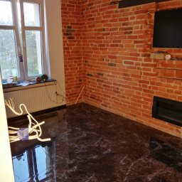 PAMIREX REMONTY I WYKONCZENIA PAWEŁ HAWRO - Pokój po remoncie z ceglaną ścianą, czarnym marmurowym podłożem, grzejnikiem pod oknem i wiszącymi kablami.