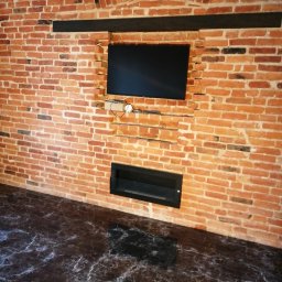 PAMIREX REMONTY I WYKONCZENIA PAWEŁ HAWRO - Ceglana ściana z wnęką na telewizor i kominek, czarna marmurowa podłoga, widoczne kable i urządzenia pod telewizorem.