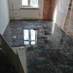 PAMIREX REMONTY I WYKONCZENIA PAWEŁ HAWRO - Układanie płytek podłogowych z ciemnego marmuru w pokoju z oknem, widoczne narzędzia i oznaczenia na podłodze, ściana zabezpieczona folią.