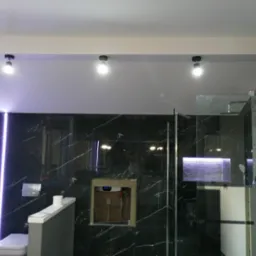 Nowoczesna łazienka z czarnymi marmurowymi płytkami, podświetlanym lustrem i kabiną prysznicową typu walk-in, z widocznym otworem w ścianie po instalacji.