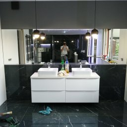 PAMIREX REMONTY I WYKONCZENIA PAWEŁ HAWRO - Nowoczesna łazienka z czarnymi płytkami imitującymi marmur, białą szafką pod umywalki i dużym lustrem, w którym odbija się osoba robiąca zdjęcie telefonem; na podłodze widoczne narzędzia i ścierka.