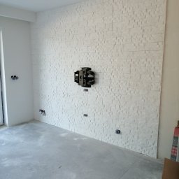 PAMIREX REMONTY I WYKONCZENIA PAWEŁ HAWRO - Ściana wykończona białym kamieniem dekoracyjnym, z zamontowanym uchwytem do telewizora, widoczne gniazdka elektryczne i surowy betonowy podkład pod podłogę.