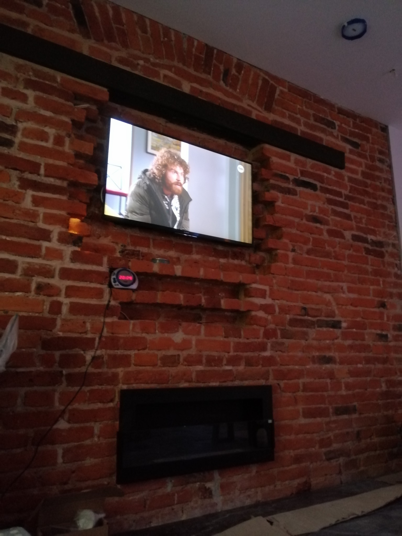 Telewizor wmurowany w ścianę z czerwonej cegły, z widocznymi śladami po kuciu i czarnymi elementami konstrukcyjnymi powyżej i poniżej ekranu, obok widoczny zegar elektroniczny.