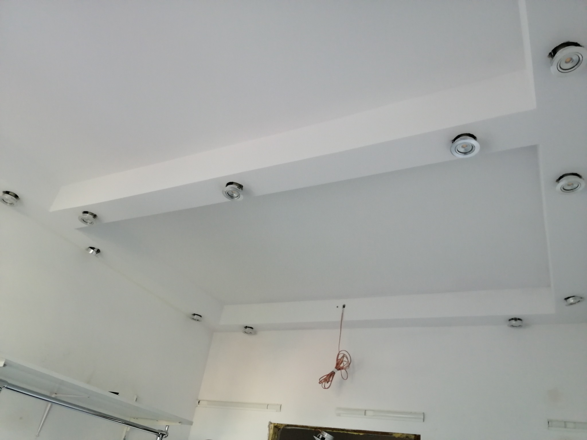 Biały sufit podwieszany z wbudowanymi okrągłymi lampami LED, widoczne przewody elektryczne zwisające ze ściany wskazujące na trwające prace instalacyjne.