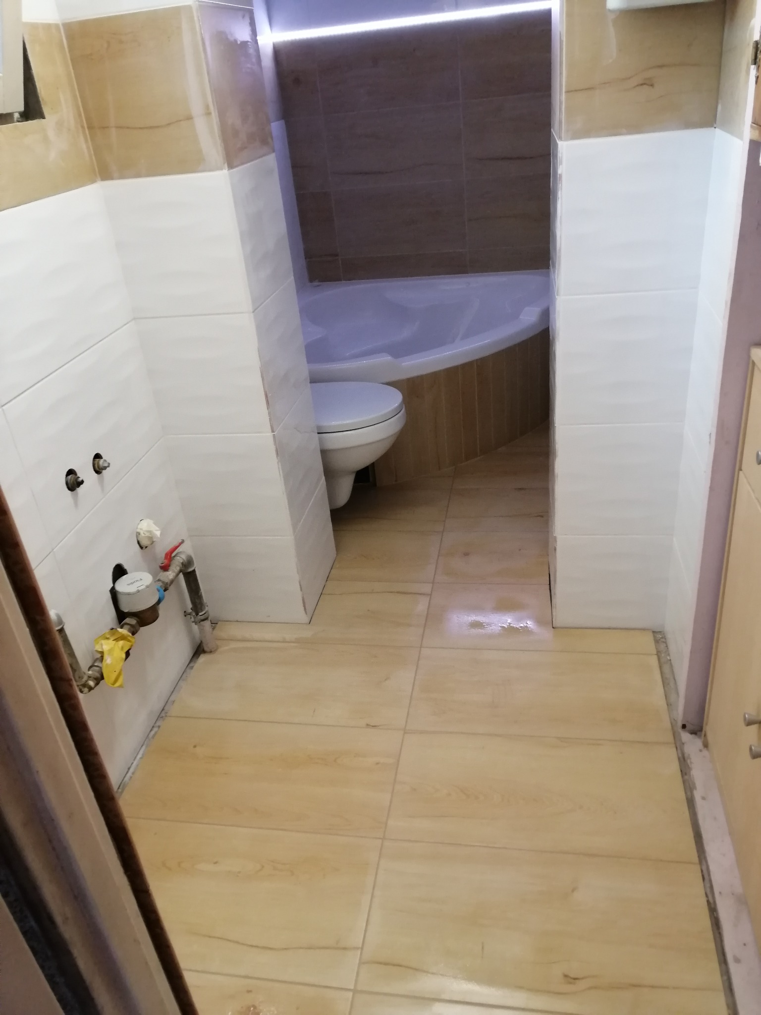 Wnętrze łazienki w trakcie prac wykończeniowych, widoczne białe płytki na ścianach, drewnopodobne płytki na podłodze, częściowo zamontowana toaleta i wanna, oraz odsłonięte rury wodociągowe z żółtą...