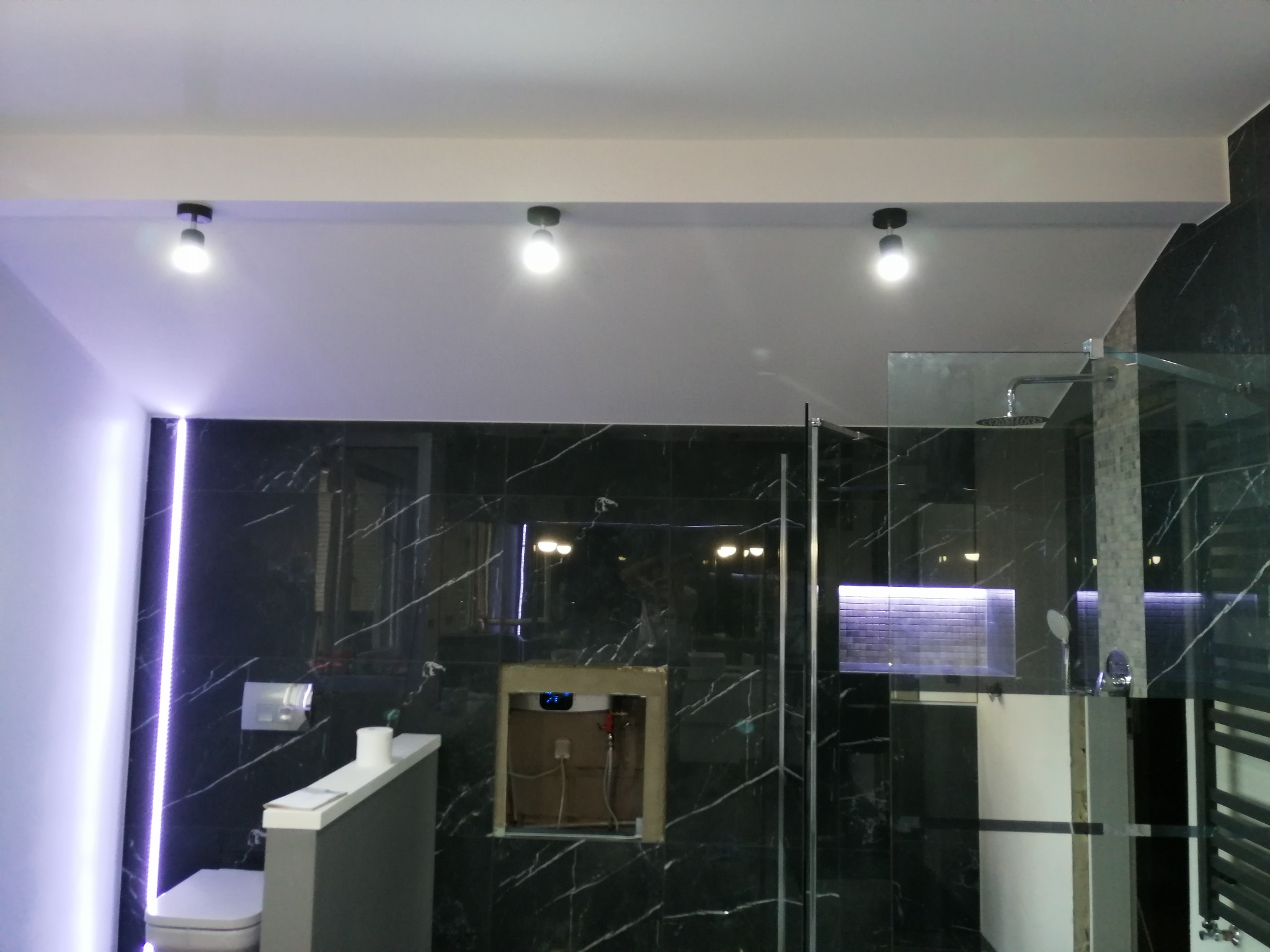 Nowoczesna łazienka z czarnymi marmurowymi płytkami, podświetlanym lustrem i kabiną prysznicową typu walk-in, z widocznym otworem w ścianie po instalacji.