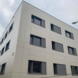 CS - Nowoczesny budynek biurowy z elewacją z paneli w kolorze beżowym i grafitowymi oknami, widok z dołu na tle pochmurnego nieba.