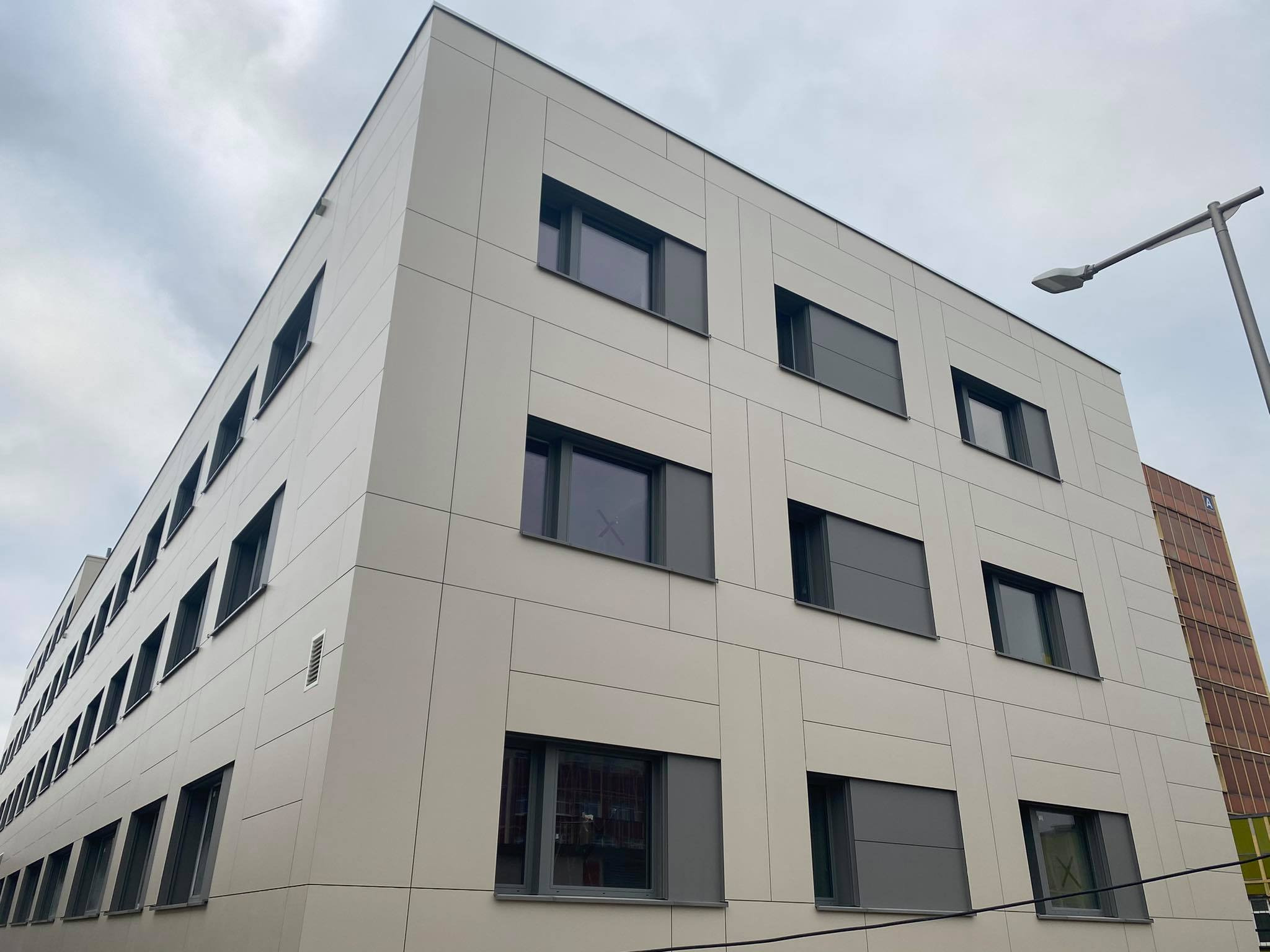 Nowoczesny budynek biurowy z elewacją z paneli w kolorze beżowym i grafitowymi oknami, widok z dołu na tle pochmurnego nieba.