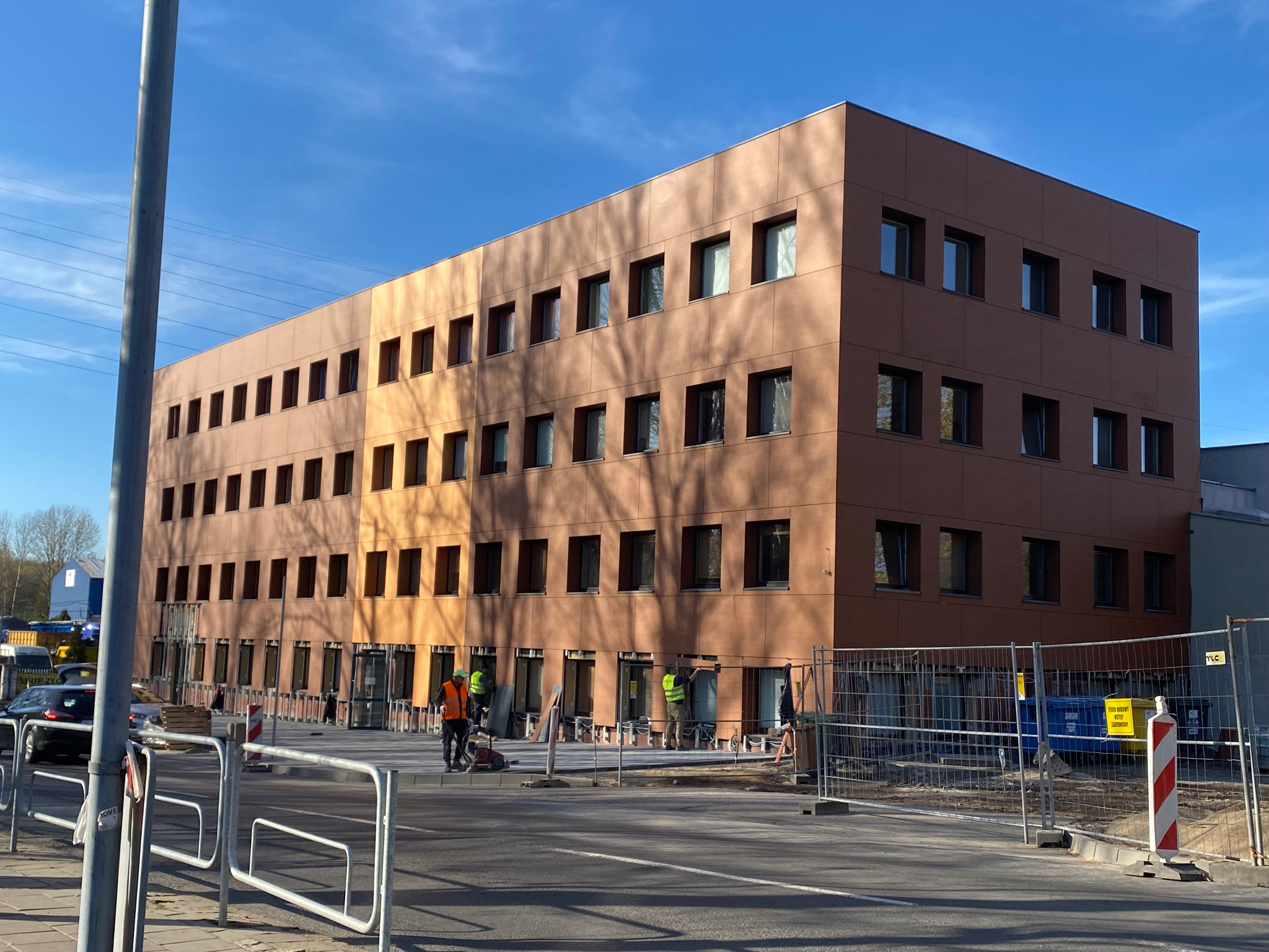 Trzykondygnacyjny budynek biurowy z brązową elewacją panelową, w trakcie montażu, widoczni pracownicy w kamizelkach odblaskowych, ogrodzenie budowy, częściowe zacienienie elewacji.