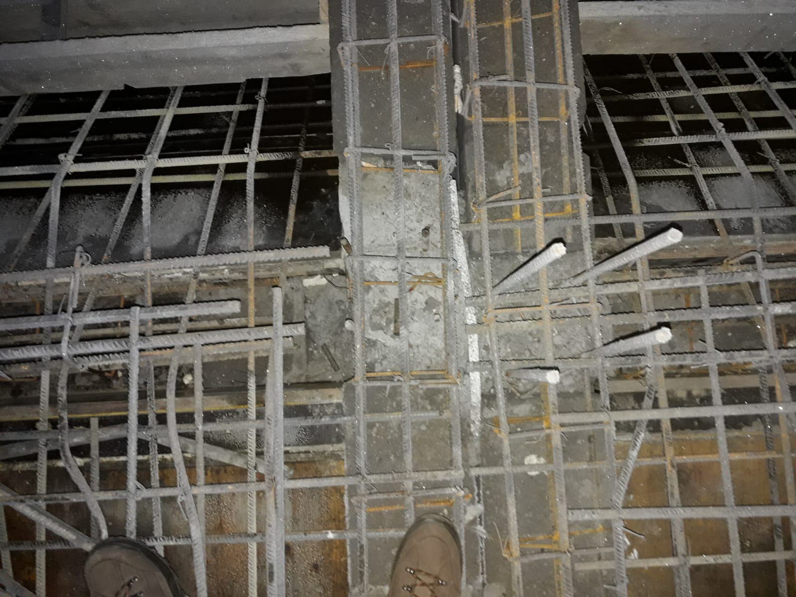 Zbrojenie kolumny i stropu przygotowane do zalania betonem, widoczne pręty zbrojeniowe połączone drutem, fragment buta w rogu kadru.