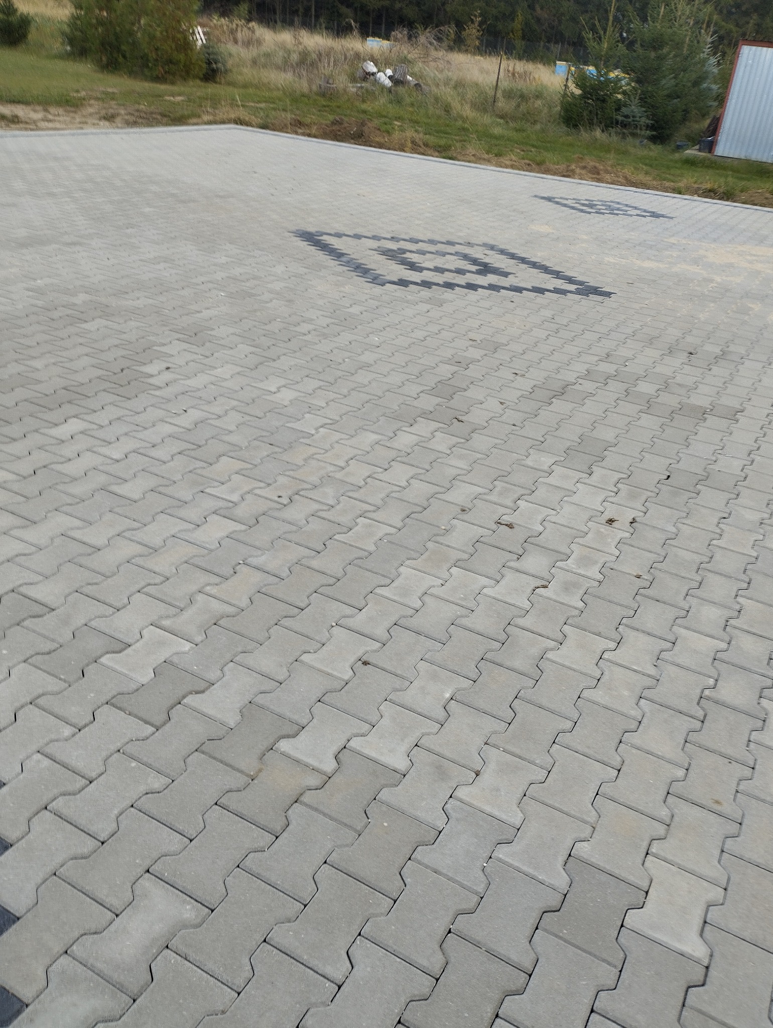 Szara kostka brukowa ułożona we wzór jodełki, z ciemnymi elementami tworzącymi geometryczny ornament w centralnej części powierzchni.