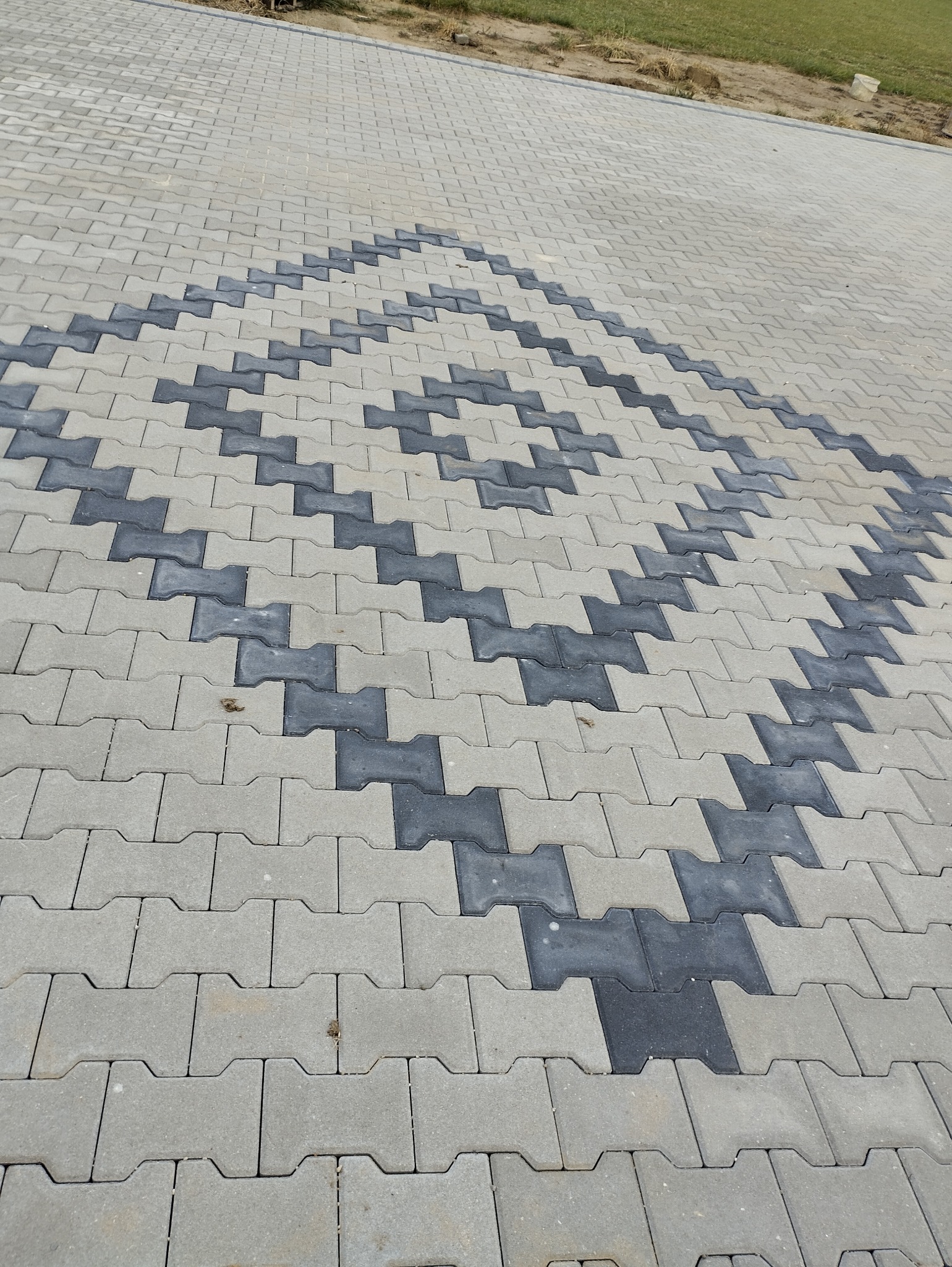 Ułożona nawierzchnia z szarej i grafitowej kostki brukowej w kształcie litery I, tworząca wzór rombu na podjeździe.
