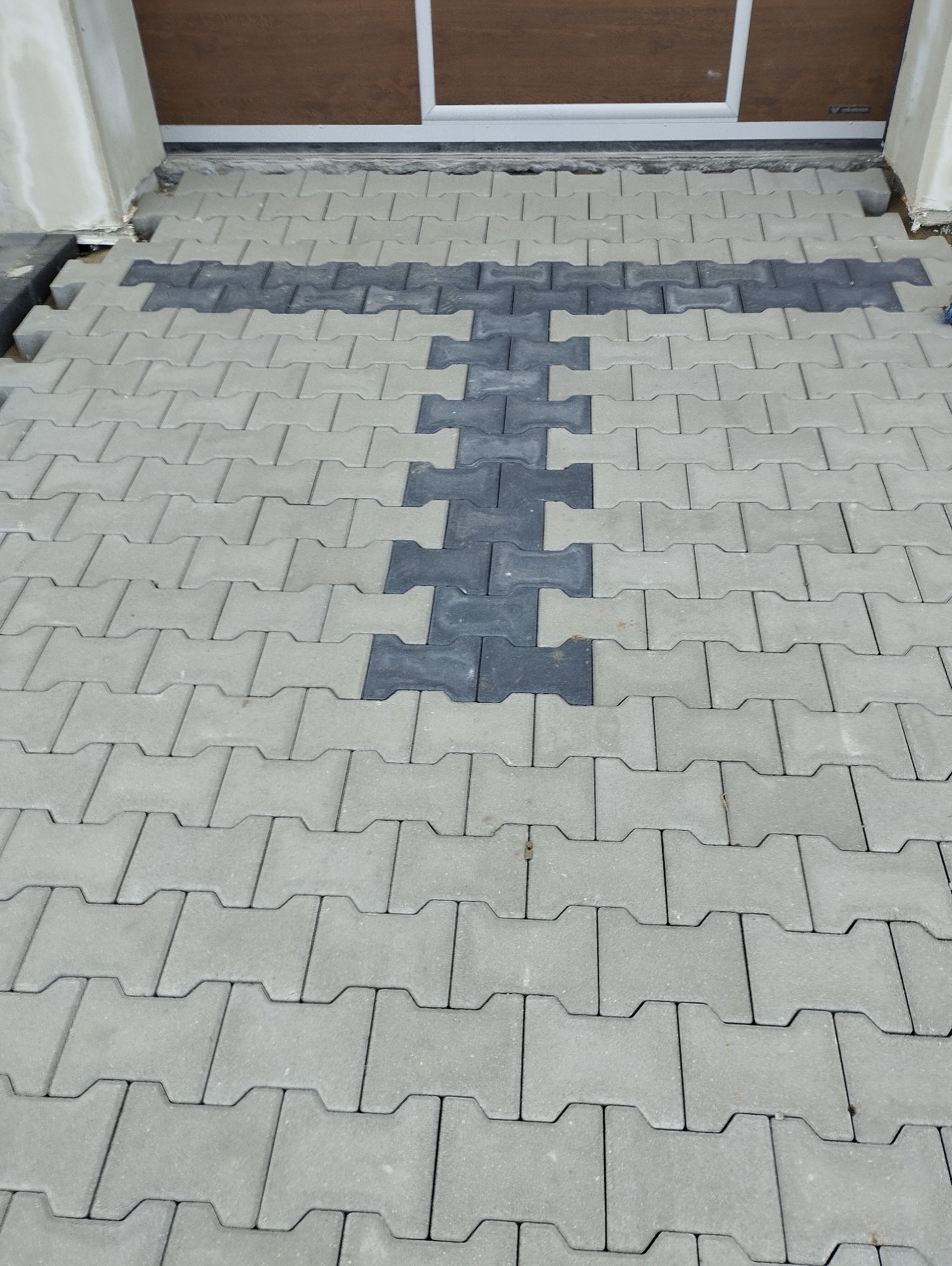 Fragment powierzchni z szarej kostki brukowej ułożonej w regularny wzór, z ciemniejszym, krzyżowym akcentem w centralnej części, tuż przed progiem drzwi wejściowych.