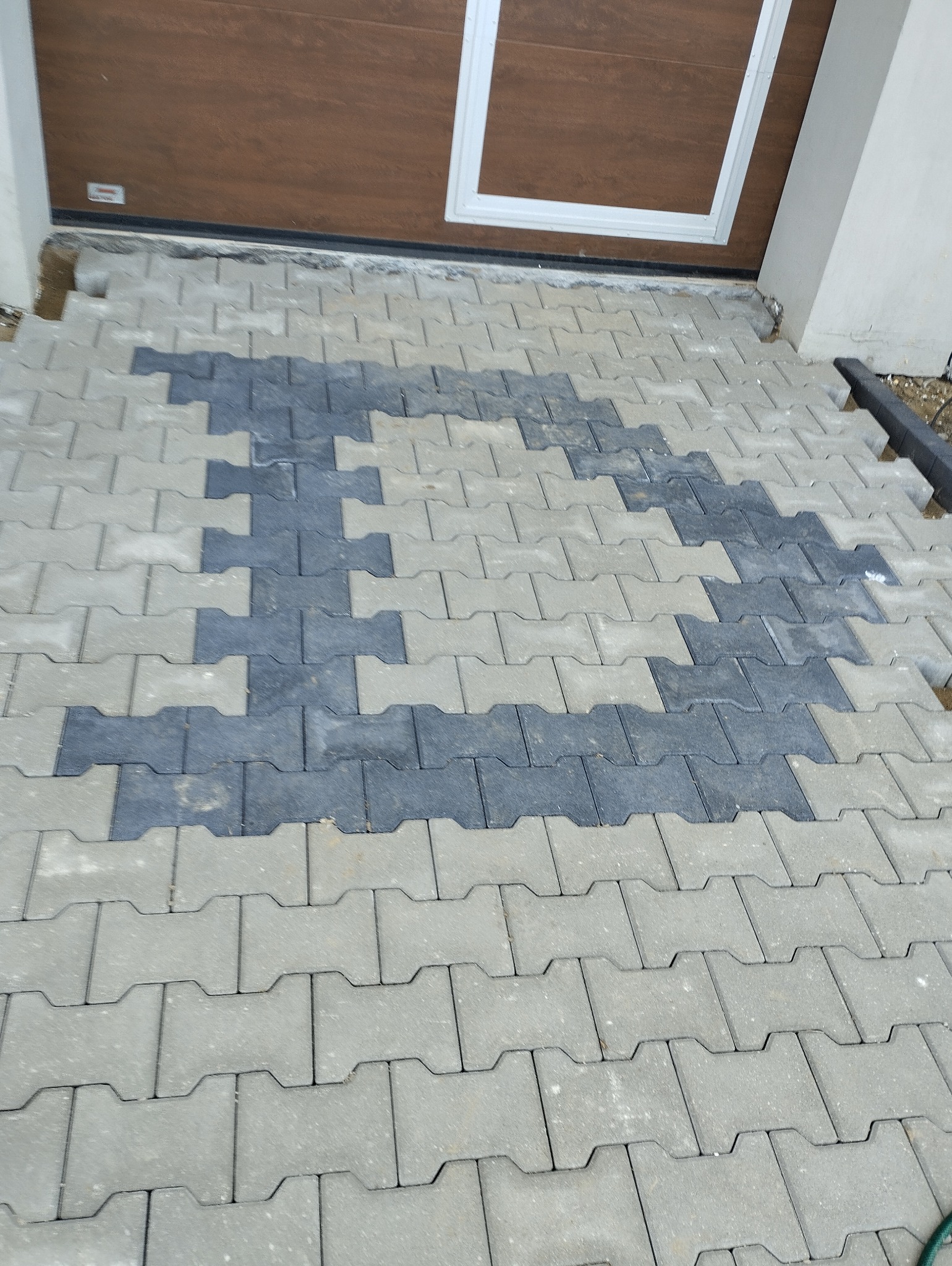 Fragment podjazdu z szarej kostki brukowej układanej w jodełkę, z wstawkami z ciemnoszarej kostki tworzącymi wzór, widoczny fragment bramy garażowej i obrzeża.