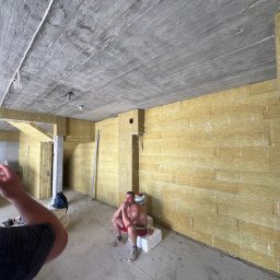 Ruslan Sobolivskyi - Wnętrze w surowym stanie z betonowym sufitem i ścianami pokrytymi izolacją z wełny mineralnej; siedzący mężczyzna w krótkich spodenkach na bloczku betonowym.