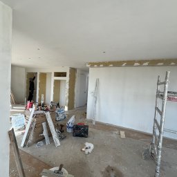 Ruslan Sobolivskyi - Wnętrze w trakcie prac wykończeniowych: ściany przygotowane do malowania, widoczne narzędzia, drabina, szpachle, worki z gładzią, pracownik w oddali szpachluje ścianę.