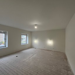 Ruslan Sobolivskyi - Pokój po świeżym malowaniu ścian na biało, widoczne niedoskonałości na podłodze z desek, okna z widokiem na budynki mieszkalne.