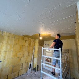 Ruslan Sobolivskyi - Mężczyzna na aluminiowym rusztowaniu, nakładający białą masę szpachlową na sufit w trakcie remontu, widoczne ściany z żółtych pustaków i wystające kable z żarówką.