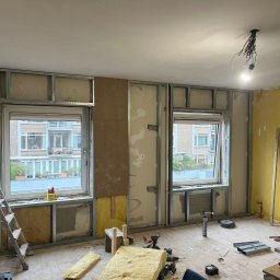 Ruslan Sobolivskyi - Pokój w trakcie remontu: widoczne metalowe profile konstrukcyjne przy oknach, częściowo ocieplone wełną mineralną, odsłonięte przewody elektryczne zwisające z sufitu, drabina oparta o ścianę...