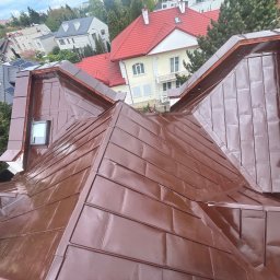 Ruslan Sobolivskyi - Błyszczący, brązowy dach z blachy trapezowej, ułożony w karo, z widoczną lukarną i fragmentami sąsiednich budynków w tle.