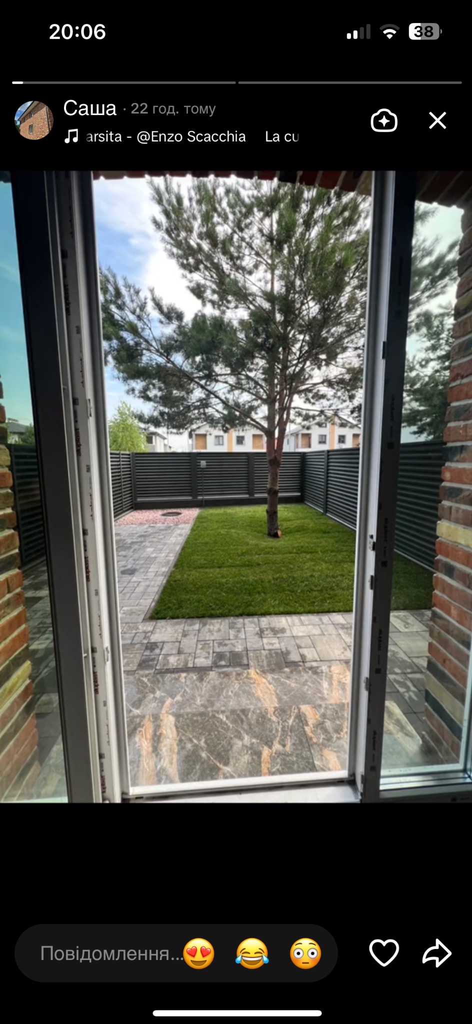 Widok na zadbany ogród z trawnikiem i drzewem przez otwarte drzwi balkonowe, z widocznymi płytami chodnikowymi i metalowym ogrodzeniem.