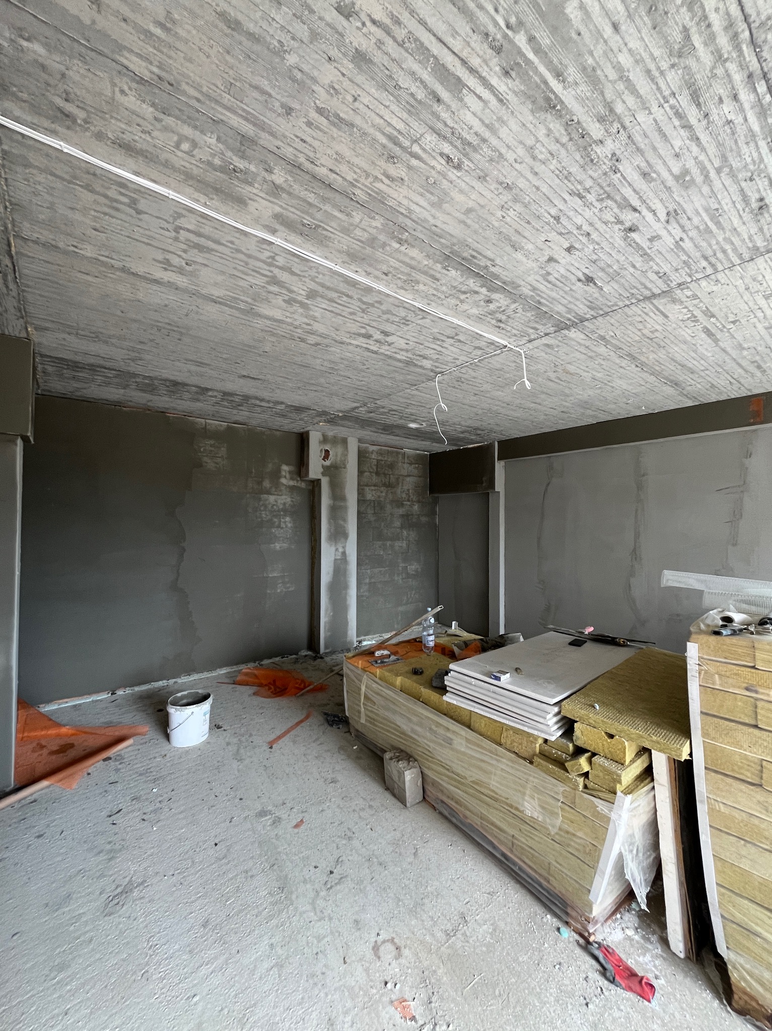 Surowy stan wnętrza w trakcie remontu: ściany po gładziowaniu, widoczny surowy betonowy strop z instalacją elektryczną, materiały budowlane przygotowane do dalszych prac.