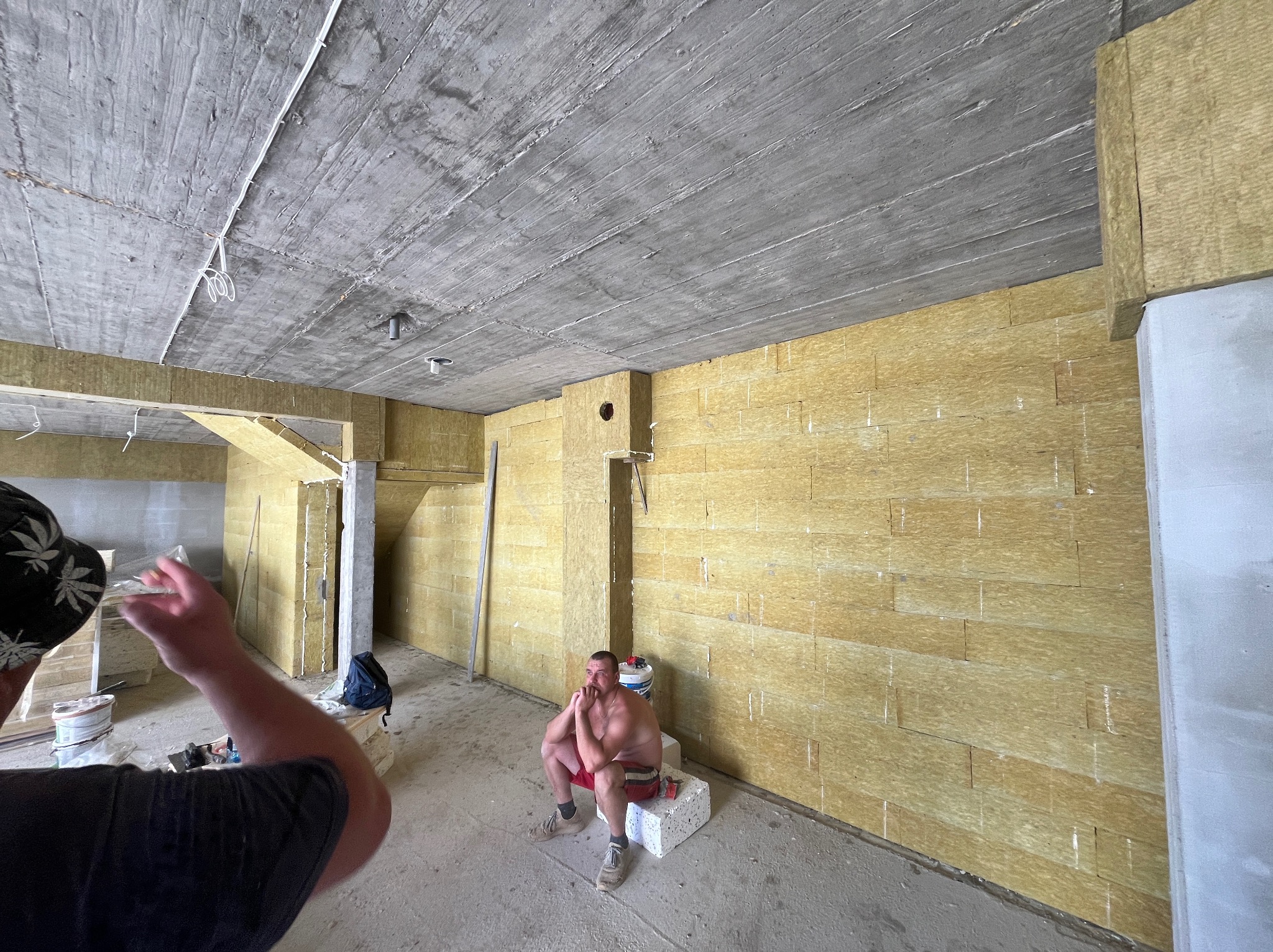 Wnętrze w surowym stanie z betonowym sufitem i ścianami pokrytymi izolacją z wełny mineralnej; siedzący mężczyzna w krótkich spodenkach na bloczku betonowym.