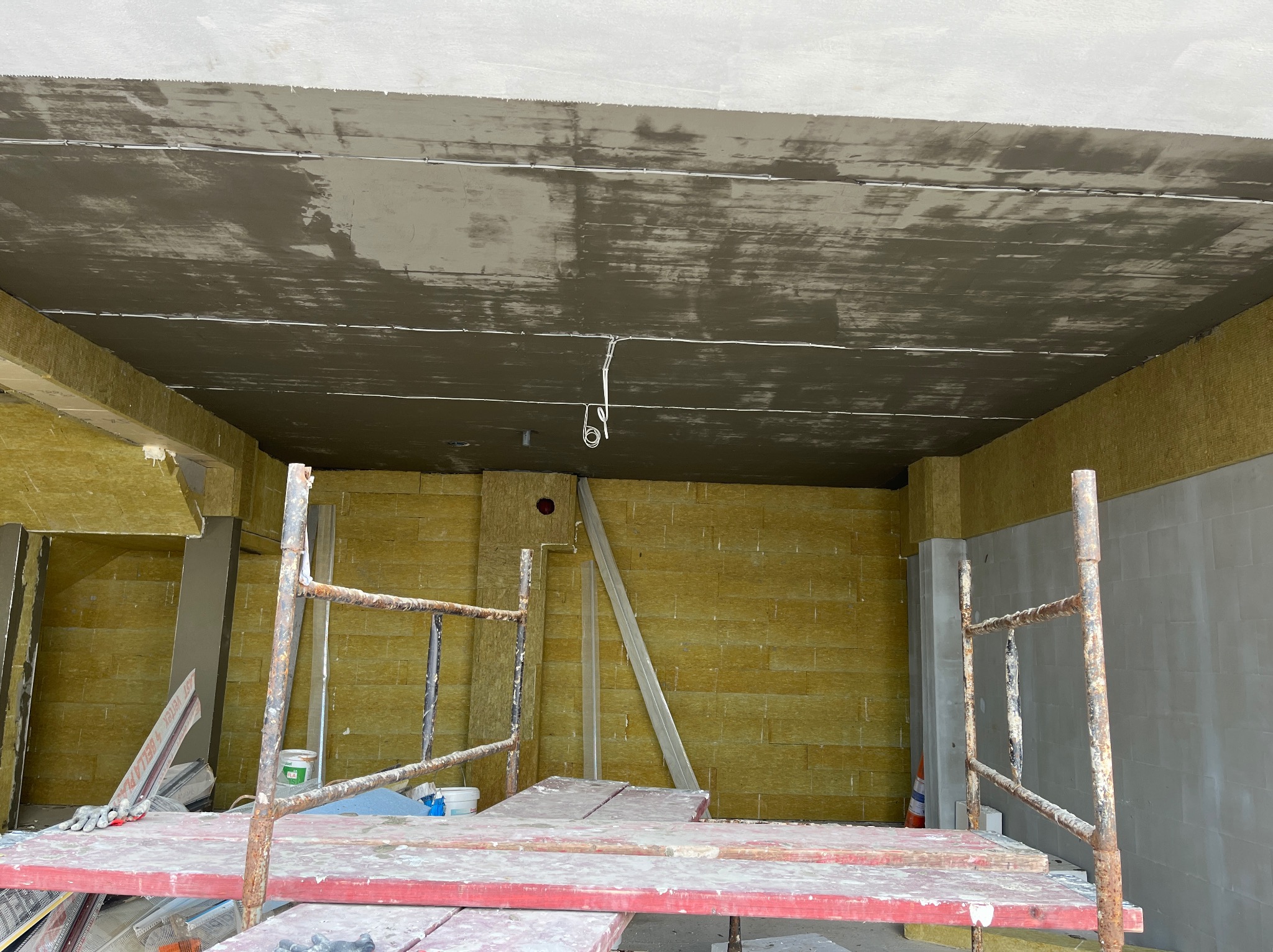 Surowy strop z widocznymi przewodami elektrycznymi w trakcie prac wykończeniowych, ściany wyłożone wełną mineralną, w tle rusztowanie malarskie.