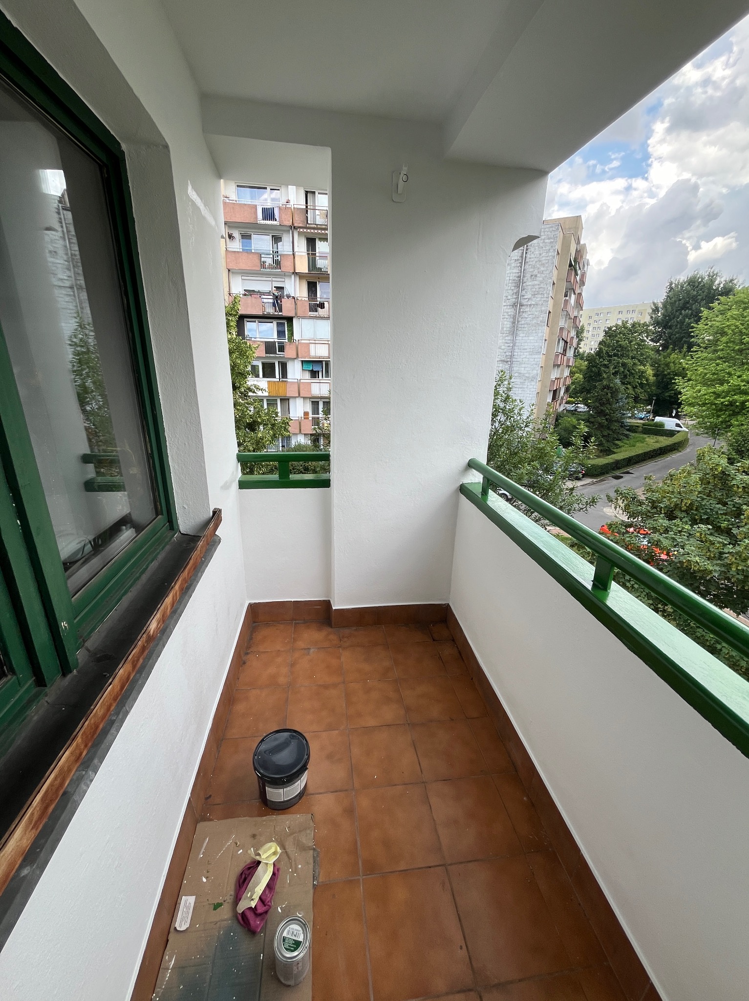 Świeżo pomalowany balkon z widokiem na budynki mieszkalne i zieleń, widoczne narzędzia malarskie: puszki z farbą, pędzel i wiadro.