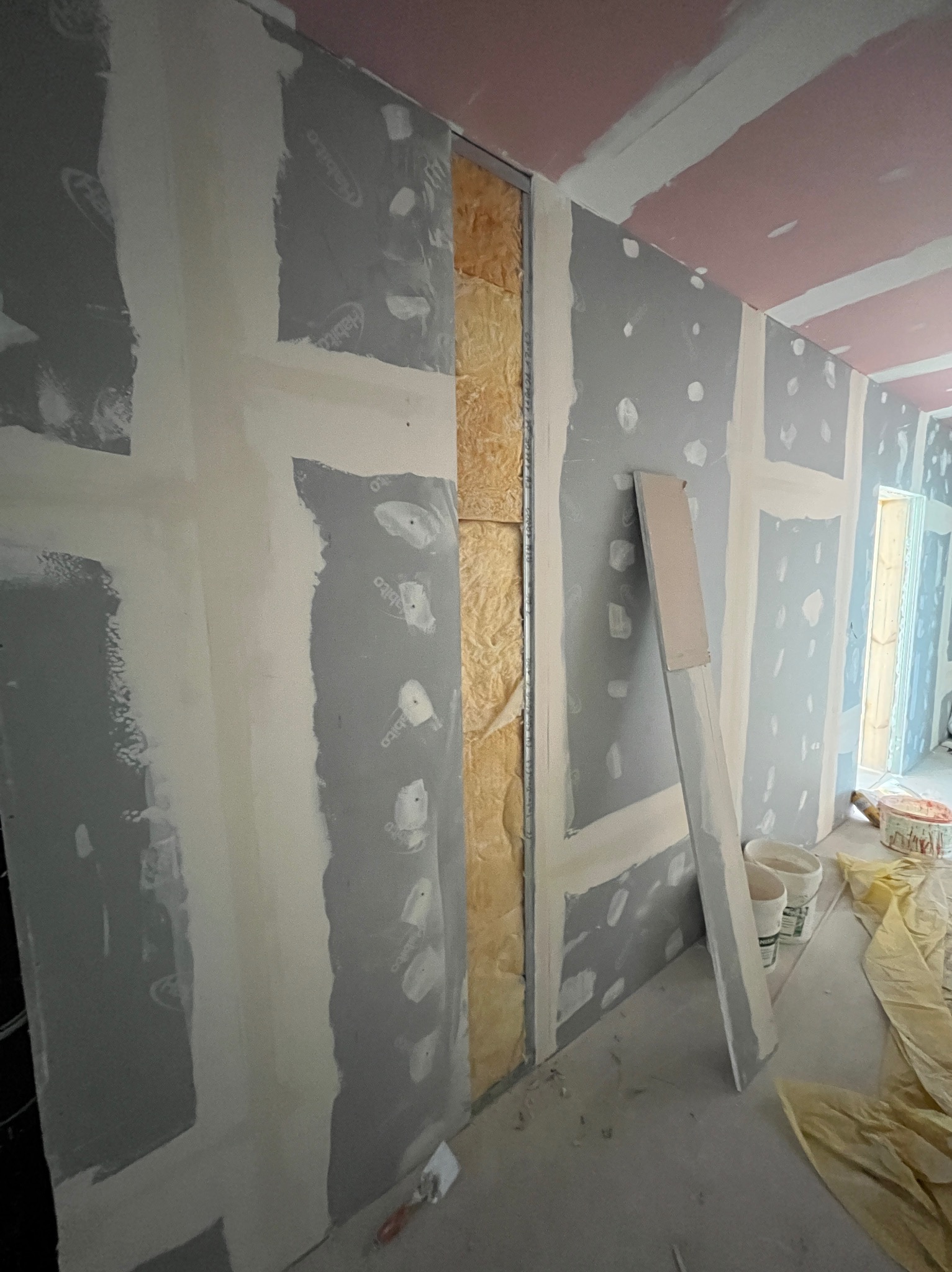 Wnętrze w trakcie montażu płyt kartonowo-gipsowych, widoczne profile, izolacja oraz wstępnie zaszpachlowane łączenia płyt na ścianach i suficie, narzędzia i wiaderka na podłodze.