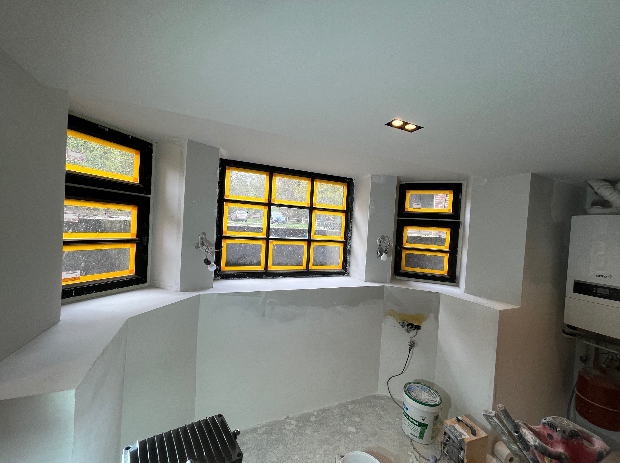 Wnętrze w Piasecznie w trakcie prac wykończeniowych: ściany po nałożeniu gładzi, okna zabezpieczone taśmą malarską, widoczne narzędzia i materiały budowlane.