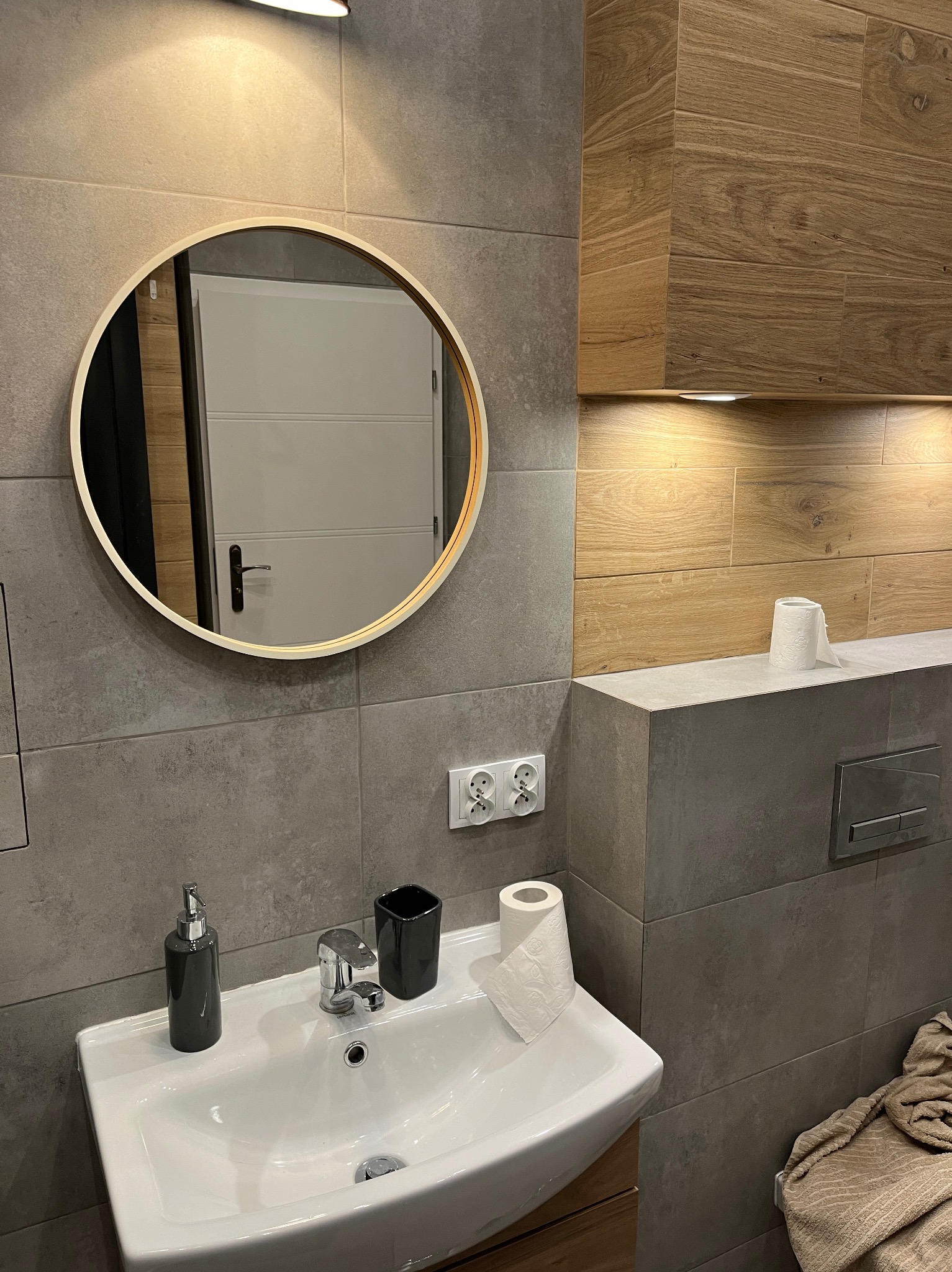 Nowoczesna łazienka z szarymi płytkami, okrągłym lustrem z podświetleniem, drewnianymi szafkami i białą umywalką z czarnymi akcesoriami