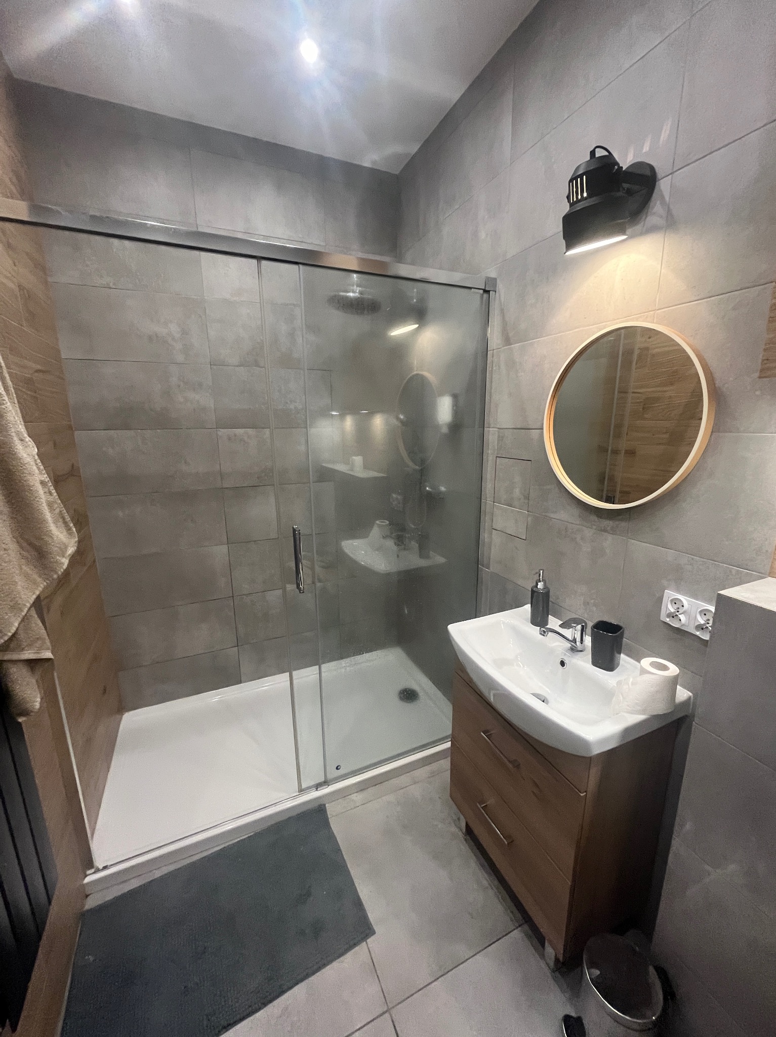 Nowoczesna łazienka z kabiną prysznicową z przesuwnymi drzwiami, umywalką nablatową na drewnianej szafce, okrągłym lustrem i szarymi płytkami imitującymi beton.