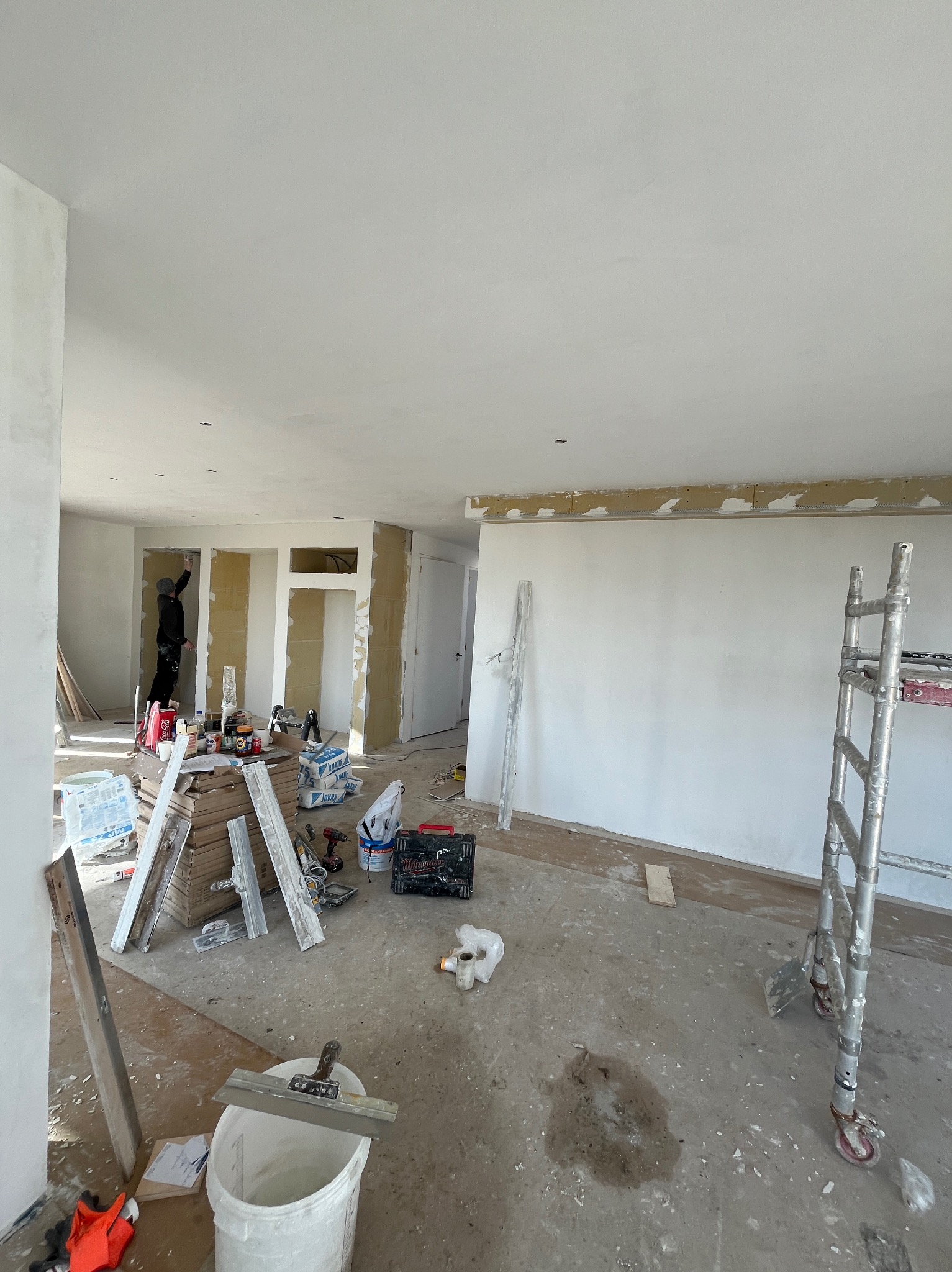 Wnętrze w trakcie prac wykończeniowych: ściany przygotowane do malowania, widoczne narzędzia, drabina, szpachle, worki z gładzią, pracownik w oddali szpachluje ścianę.