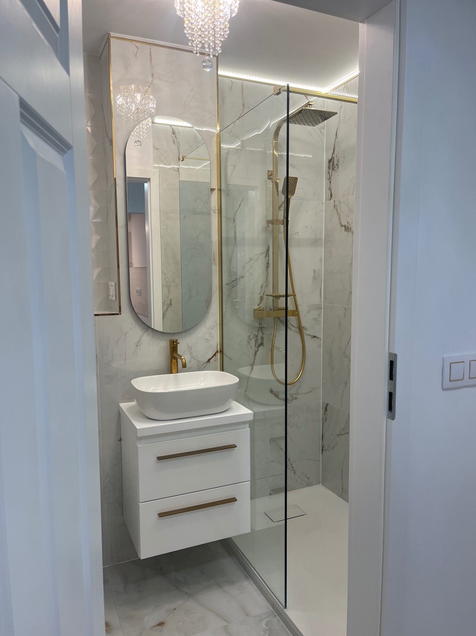 Elegancka łazienka z kabiną prysznicową typu walk-in, umywalką nablatową na szafce z szufladami i lustrem w złotej ramie. Marmurowe płytki i oświetlenie LED.