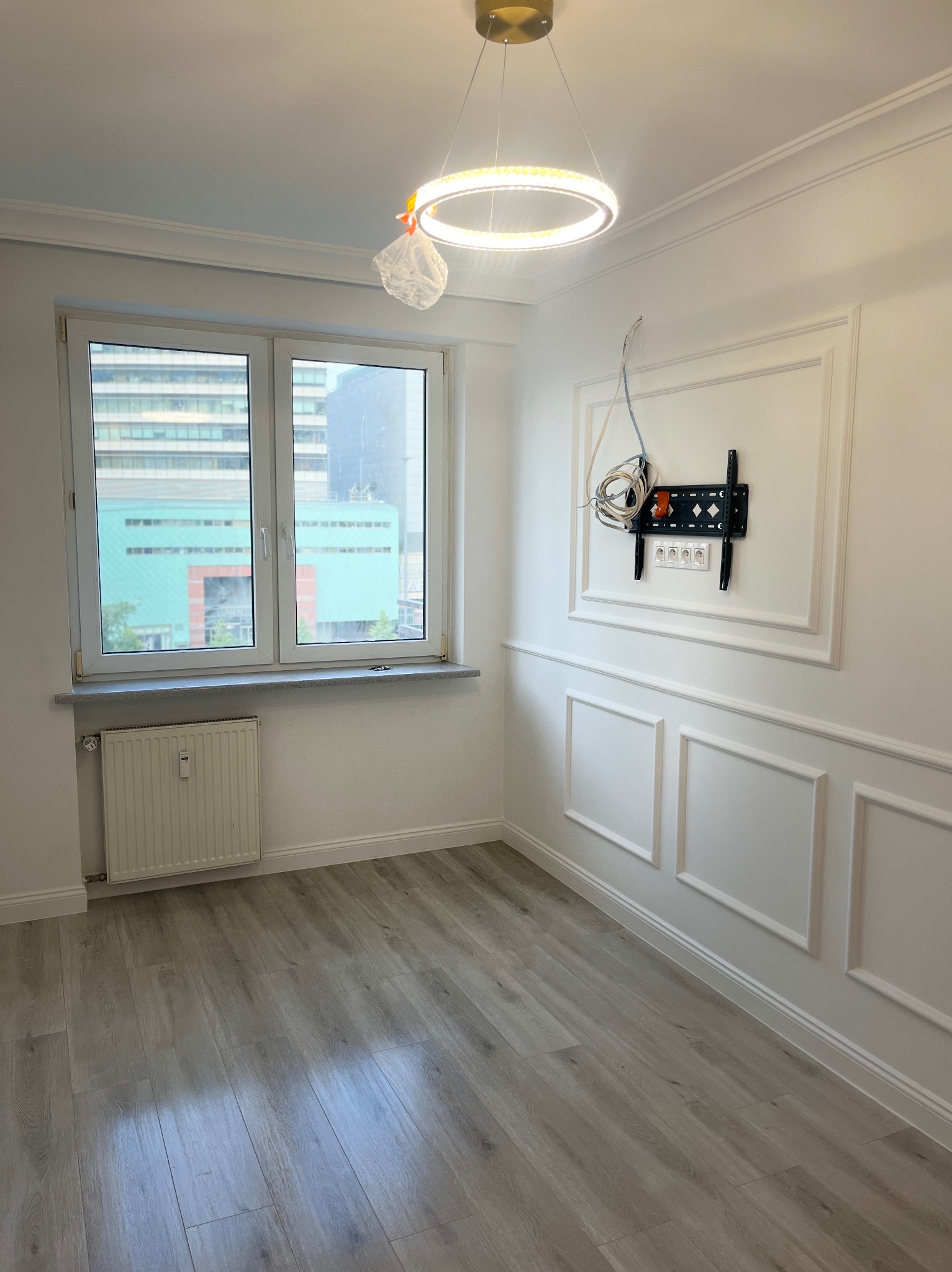 Pusty pokój po remoncie: szare panele, białe ściany z dekoracyjnymi listwami, okno z widokiem na miasto, grzejnik, nowoczesna lampa, uchwyt na TV z kablami.