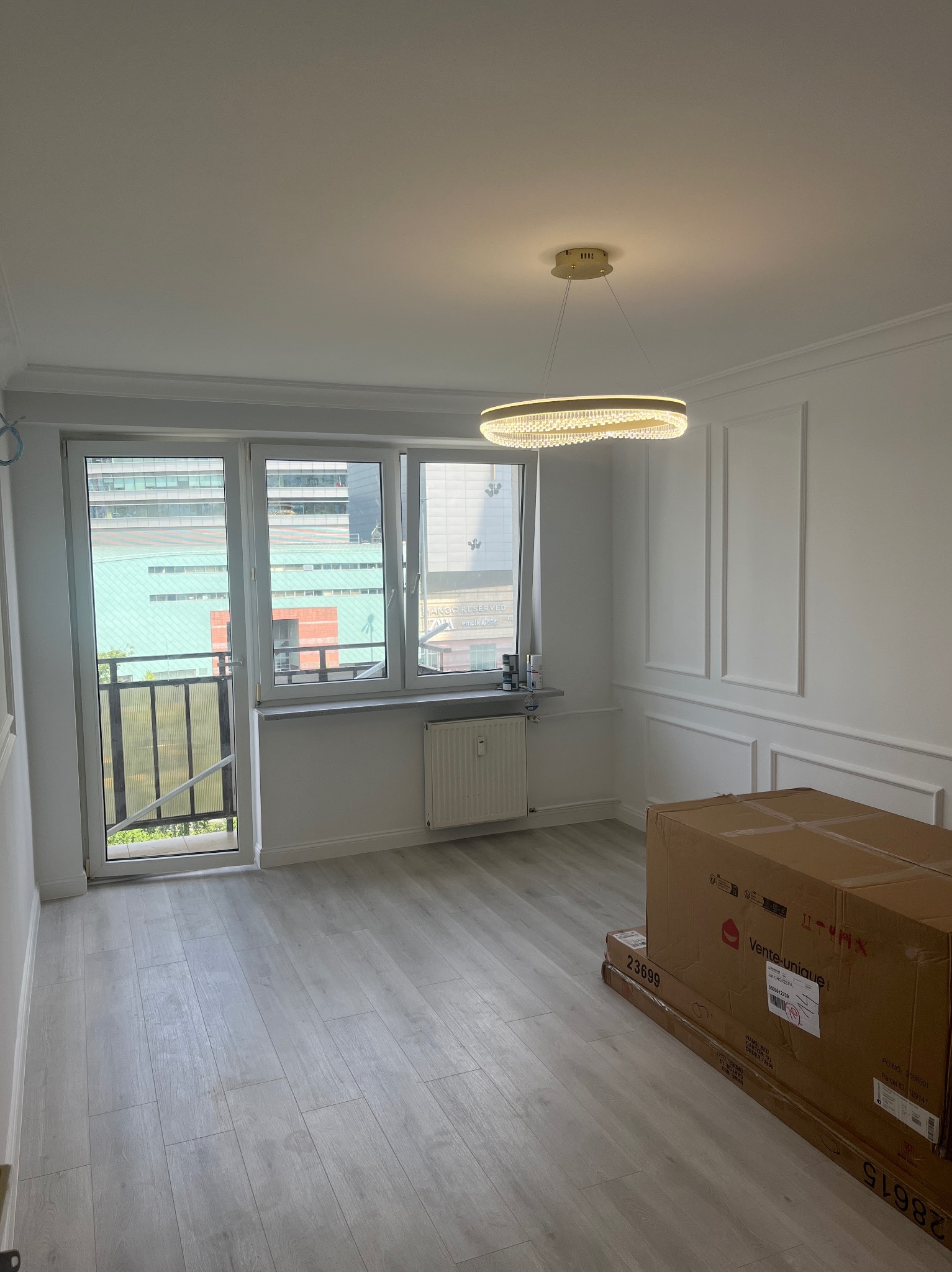 Jasne wnętrze pokoju po remoncie: białe ściany z dekoracyjnymi panelami, okno z balkonem, nowoczesna lampa i kartony na podłodze. Widok na budynek z zewnątrz.