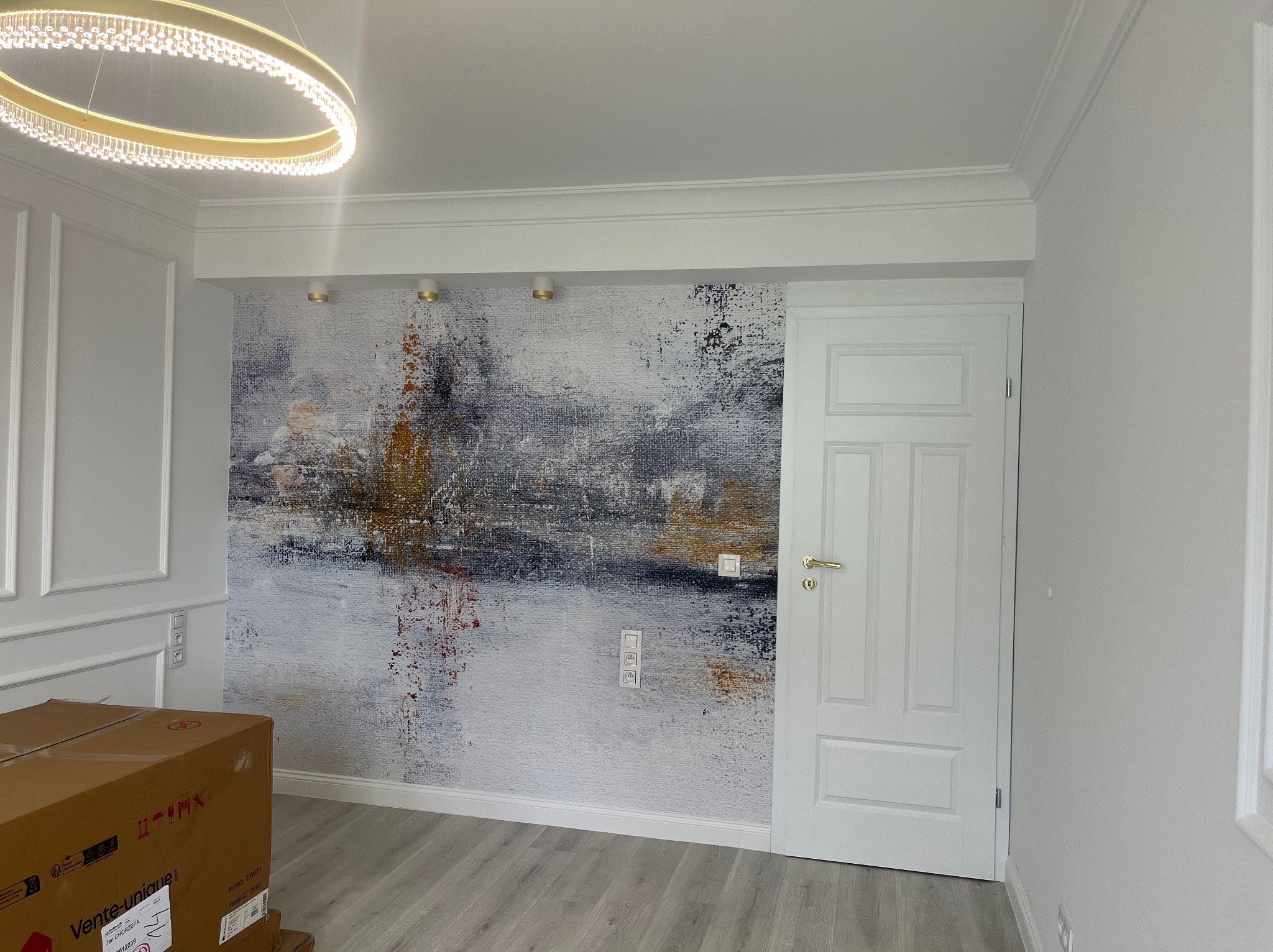 Wnętrze po odświeżeniu: białe ściany z dekoracyjną tapetą w abstrakcyjne wzory, białe listwy przypodłogowe i sufitowe, nowoczesna lampa. Częściowo widoczny karton.