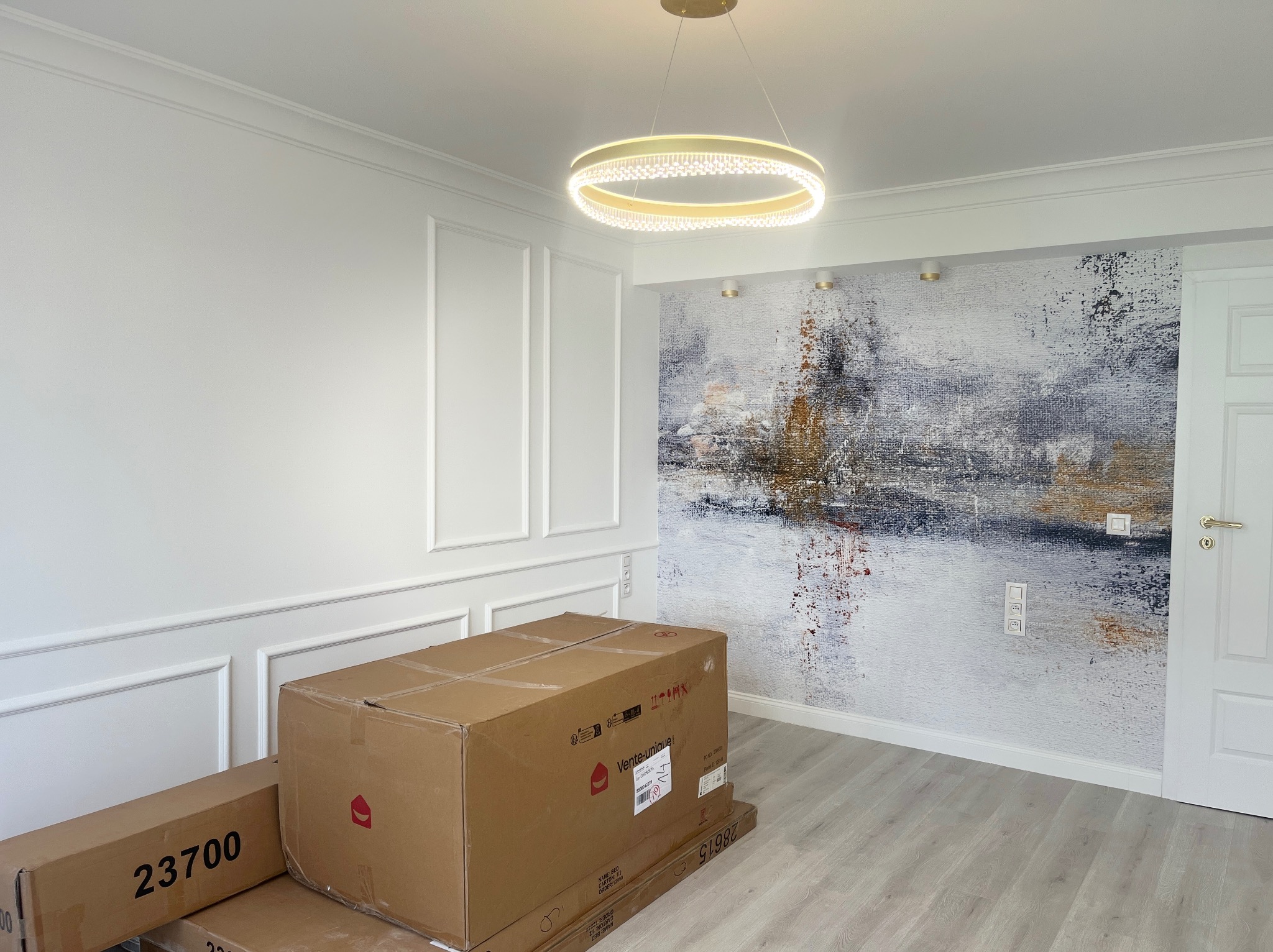 Pokój po remoncie: białe ściany z dekoracyjnymi listwami, abstrakcyjna tapeta, nowoczesna lampa, podłoga w panelach i kartony z nowymi meblami.