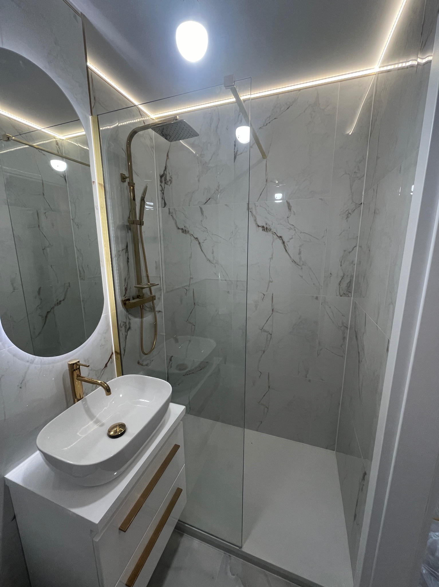 Nowoczesna łazienka z kabiną prysznicową typu walk-in, umywalką nablatową na szafce i podświetlanym lustrem w złotej ramie. Marmurowe płytki na ścianach i oświetlenie LED.