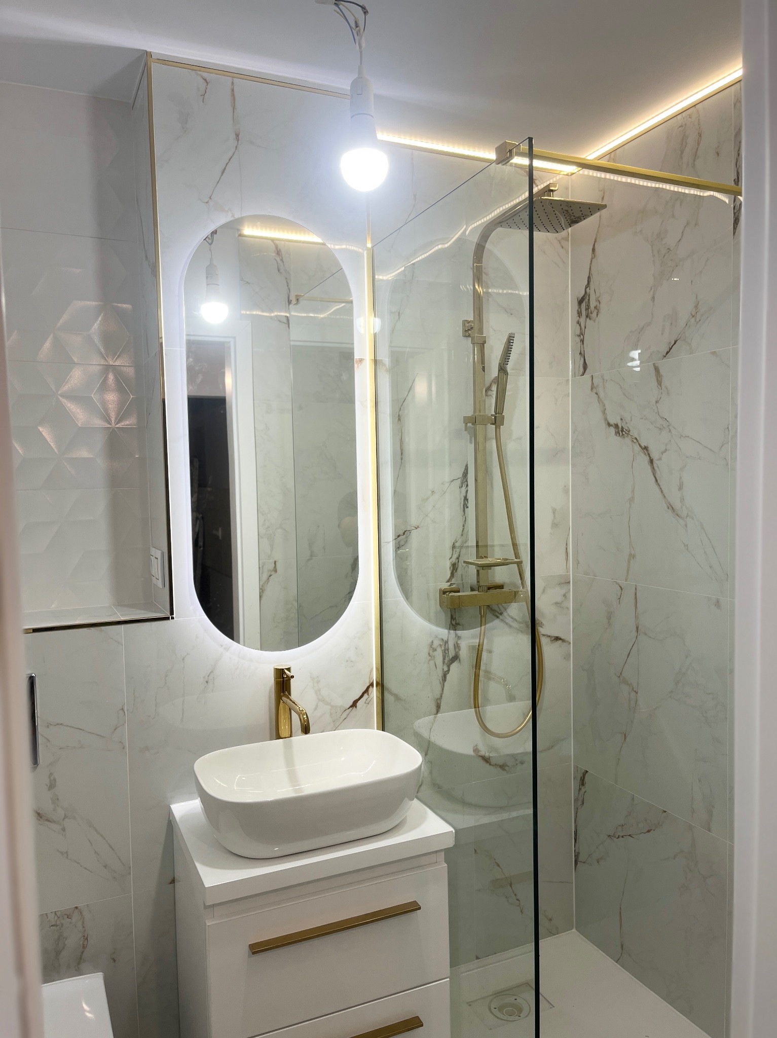 Biała łazienka z marmurowymi płytkami, złotą armaturą i podświetlanym lustrem. Kabina prysznicowa ze szklaną ścianką. Nowoczesny design.